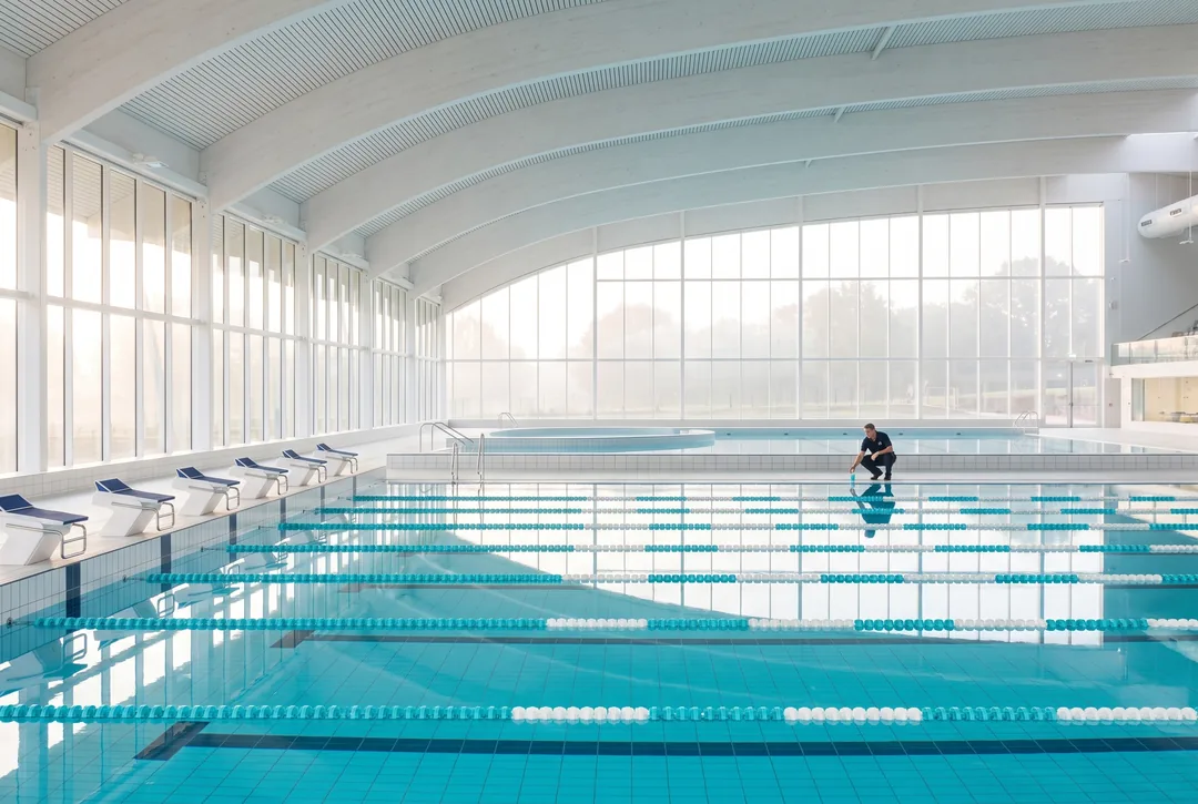 A bright indoor municipal aquatic center with a 50-meter competition pool and a leisure pool beyond, a pool technician taking a water sample far along the deck.